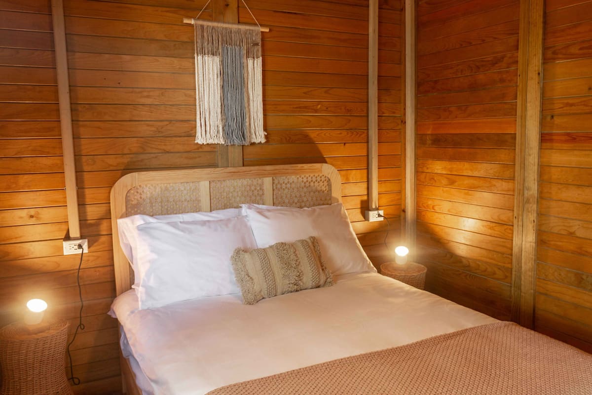 Ein Schlafzimmer einer Holzhütte mit einem Bett, einer Lampenschirm aus Makramee und einem gewebten Korb.