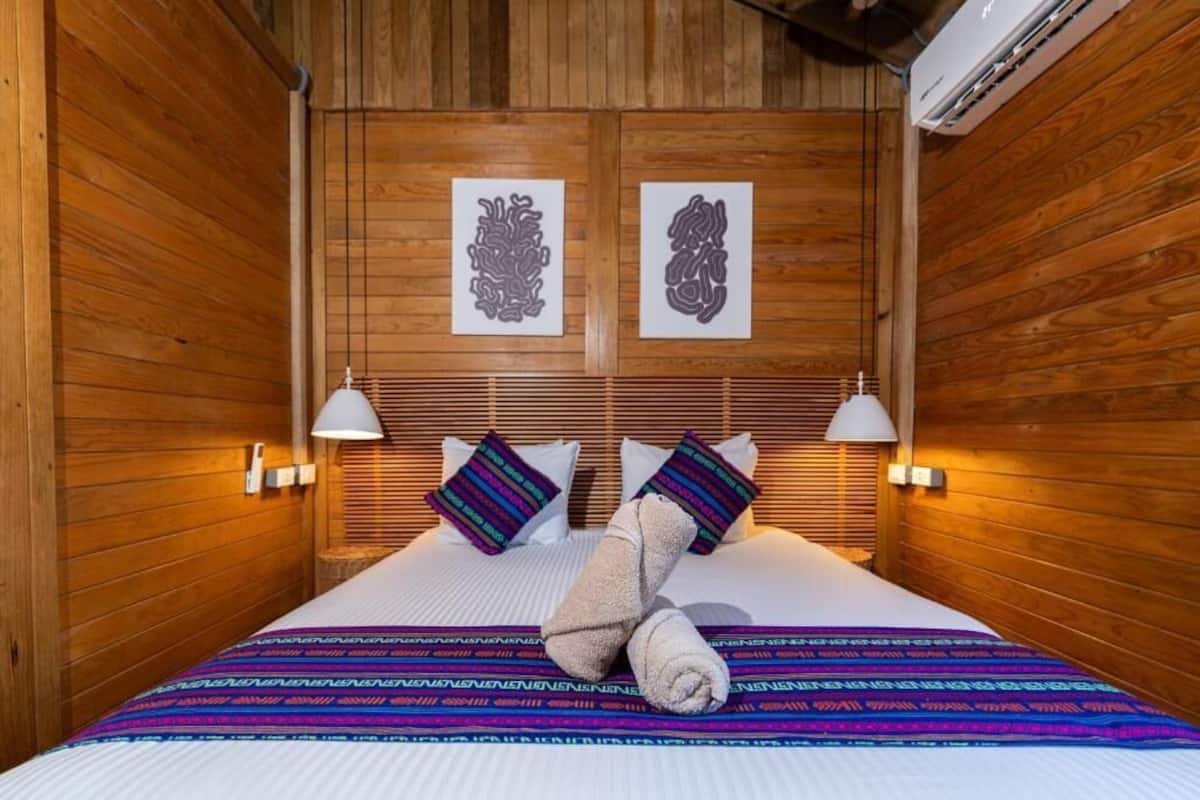 Ein Zimmer mit Holzwänden, einem Bett, zwei Wandlampen und zwei gerahmten Kunstwerken über dem Bett.