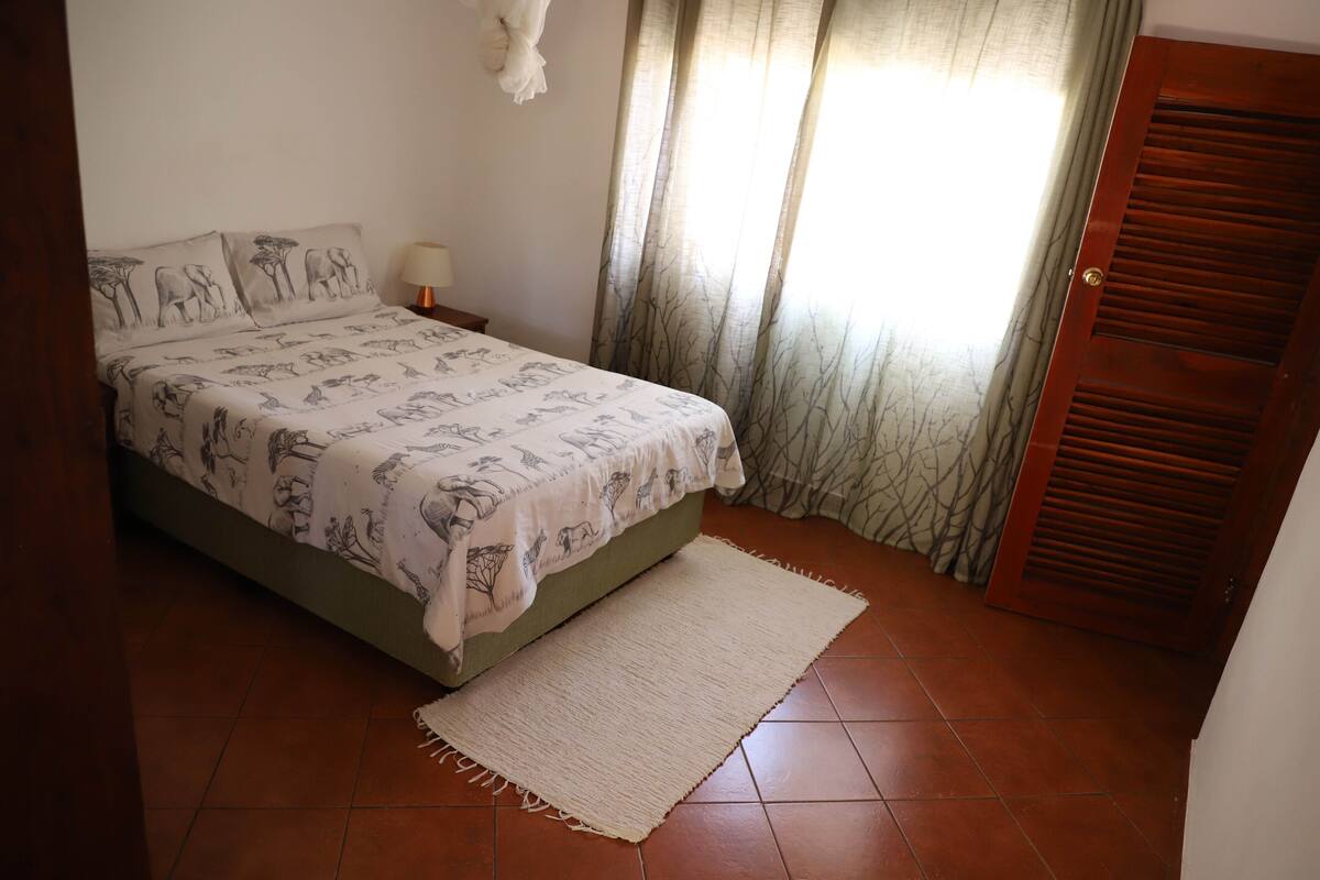 A bedroom with a bed, a nightstand, a lamp, a door, and curtains.