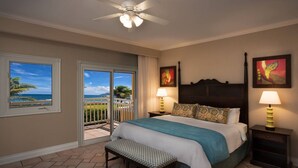 A hotel room with a bed, two bedside lamps, a ceiling fan, a bench, a window with a view of the ocean, and two paintings on the wall.