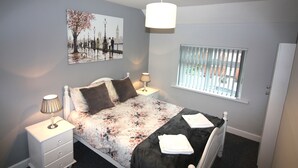 Un dormitorio con cama, mesitas de noche, ventana con persianas, lámpara colgante y un cuadro de paisaje urbano en la pared.