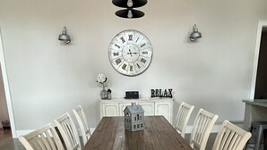 Een eetkamer met een houten tafel, witte stoelen, een wandklok en een bord met de tekst 'RELAX'.
