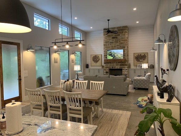 A modern kitchen with a stone fireplace, dining area with white chairs, and a large window.