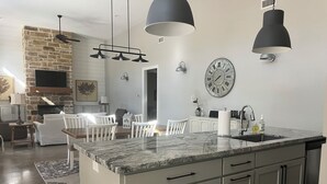 Een moderne keuken met een marmeren aanrechtblad, grijze kastjes en een inbouw kookeiland. Er hangt een grote klok aan de muur en hanglampen boven het eiland.