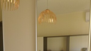 Habitación de hotel con un espejo grande que refleja una cama con sábanas blancas, un almohadón verde y una toalla doblada al lado. Se observa una lámpara de noche y un equipo de aire acondicionado instalado en la pared.