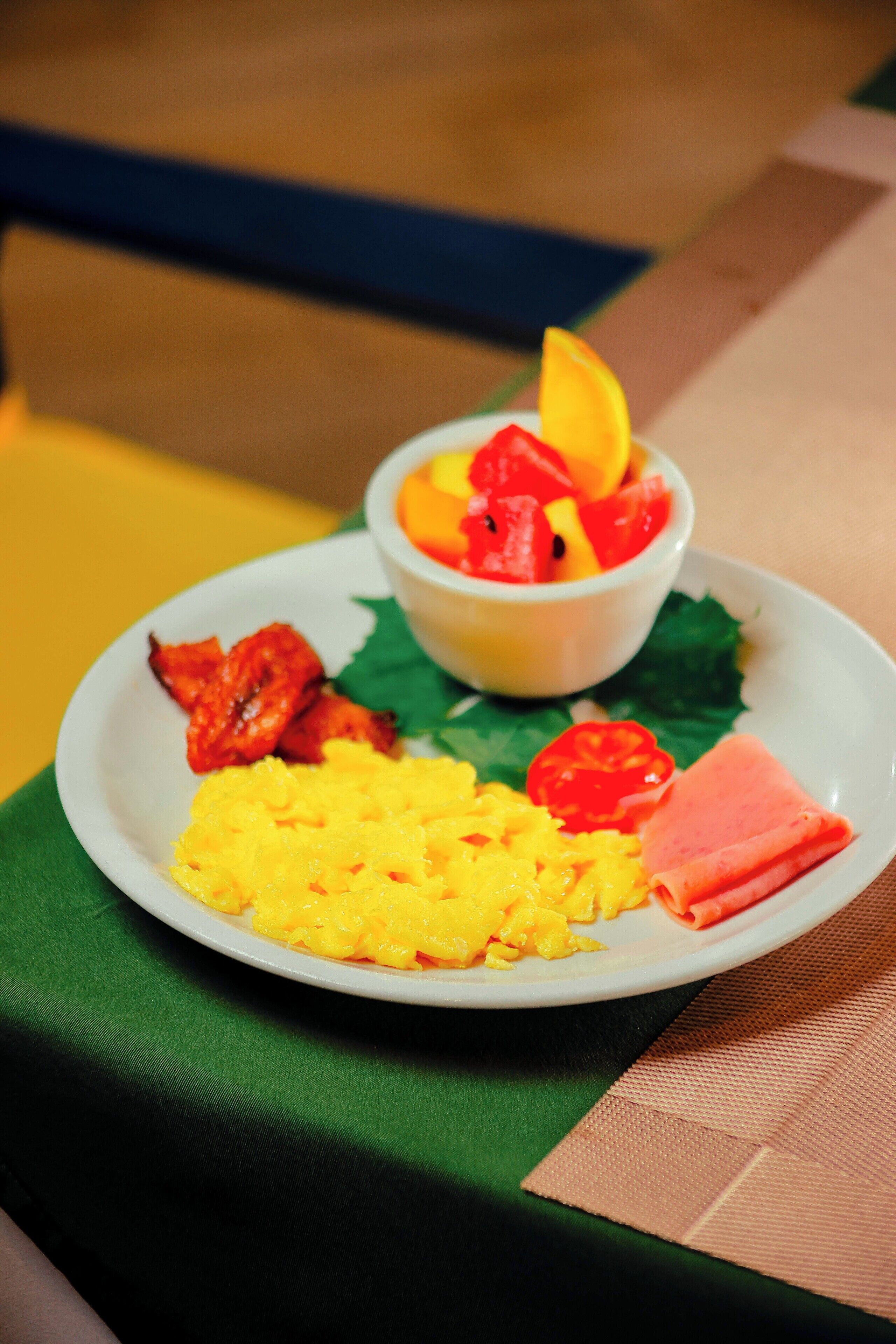 A plate of food with scrambled eggs, bacon, ham, and a fruit bowl containing watermelon and orange slices.