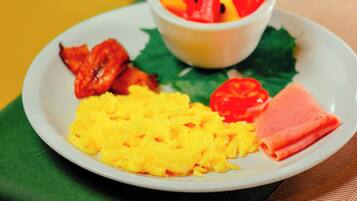 A plate of food with scrambled eggs, bacon, ham, and a fruit bowl containing watermelon and orange slices.