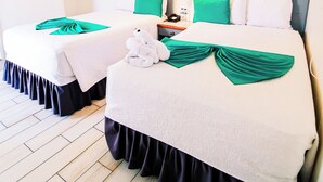 A hotel room with two beds, each with a white bedspread and green pillows. There are two bedside tables with lamps, a ceiling fan, and a wall-mounted shelf with pictures. - Hotel Casa Turquesa (Flores)