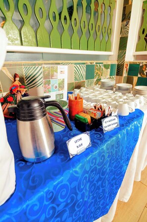 A buffet table with a blue tablecloth, a stainless steel coffee pot, and labeled tea and sugar dispensers. - Hotel Casa Turquesa (Flores)