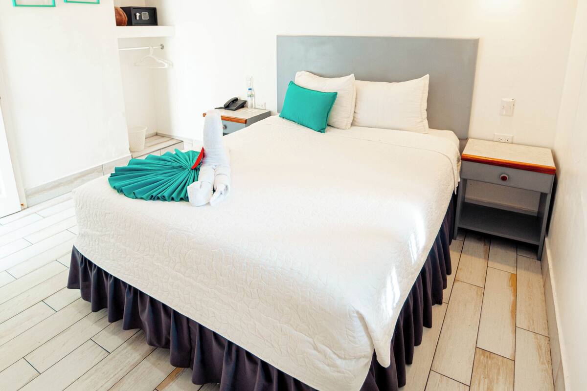 A neatly made bed with a white duvet, teal pillow, and a dark skirted bed skirt. There is a bedside table with a lamp and a nightstand with a drawer. The room has white walls, a wooden floor, and a ceiling with exposed beams.