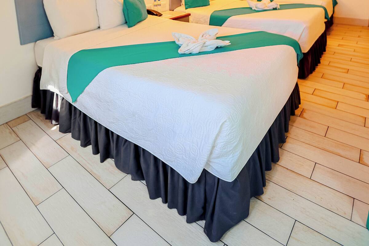 A hotel room with four beds, each with a white bedspread and teal pillows, and a wooden floor.