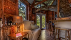 A cozy log cabin interior with a wooden sofa, a table with a lamp, and a view of the outdoors through large windows. - Savannah's Meadow (Celeste)