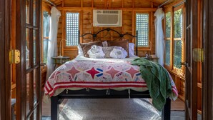 A wooden cabin bedroom with a bed, two windows, an air conditioning unit, and a chandelier. - Savannah's Meadow (Celeste)