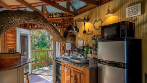 A rustic kitchen with wooden beams, a large tree trunk arching over the counter, and a view of the outdoors. - Savannah's Meadow (Celeste)