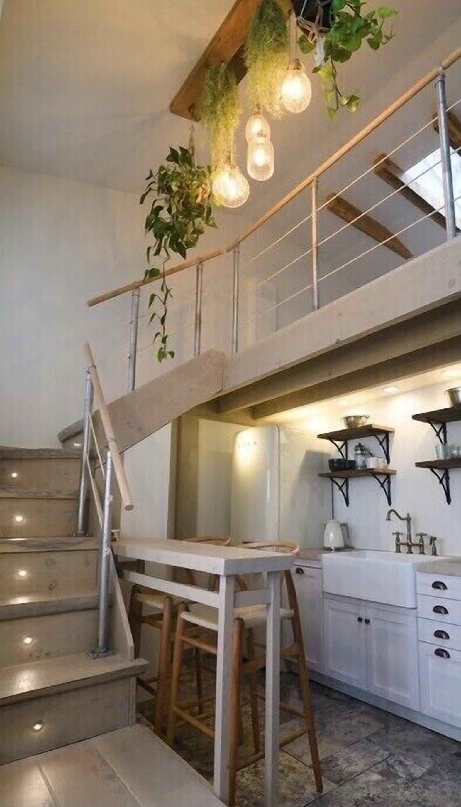 Un escalier avec une chaise en bois et une table, un coin cuisine avec des placards et un évier, et des plantes suspendues avec des lumières.