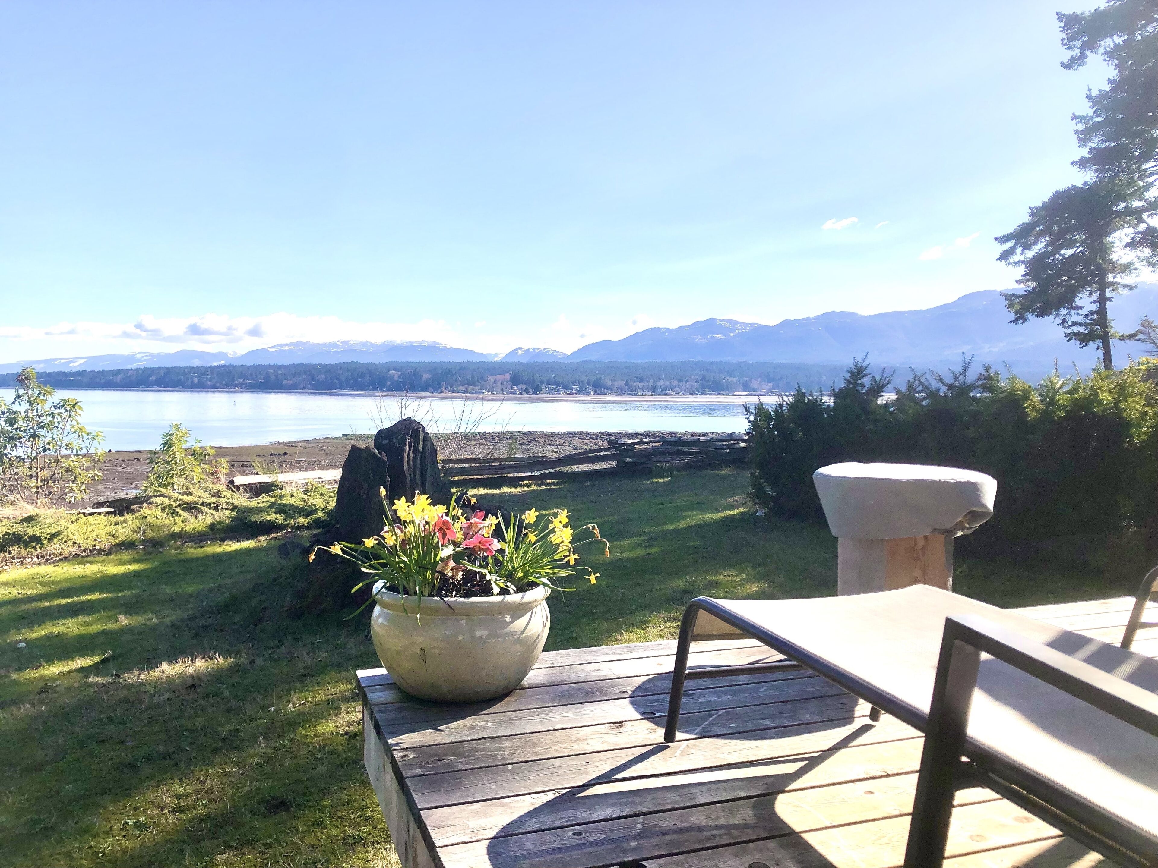 A wooden deck with a potted plant and a stone planter, overlooking a lake with mountains in the background.