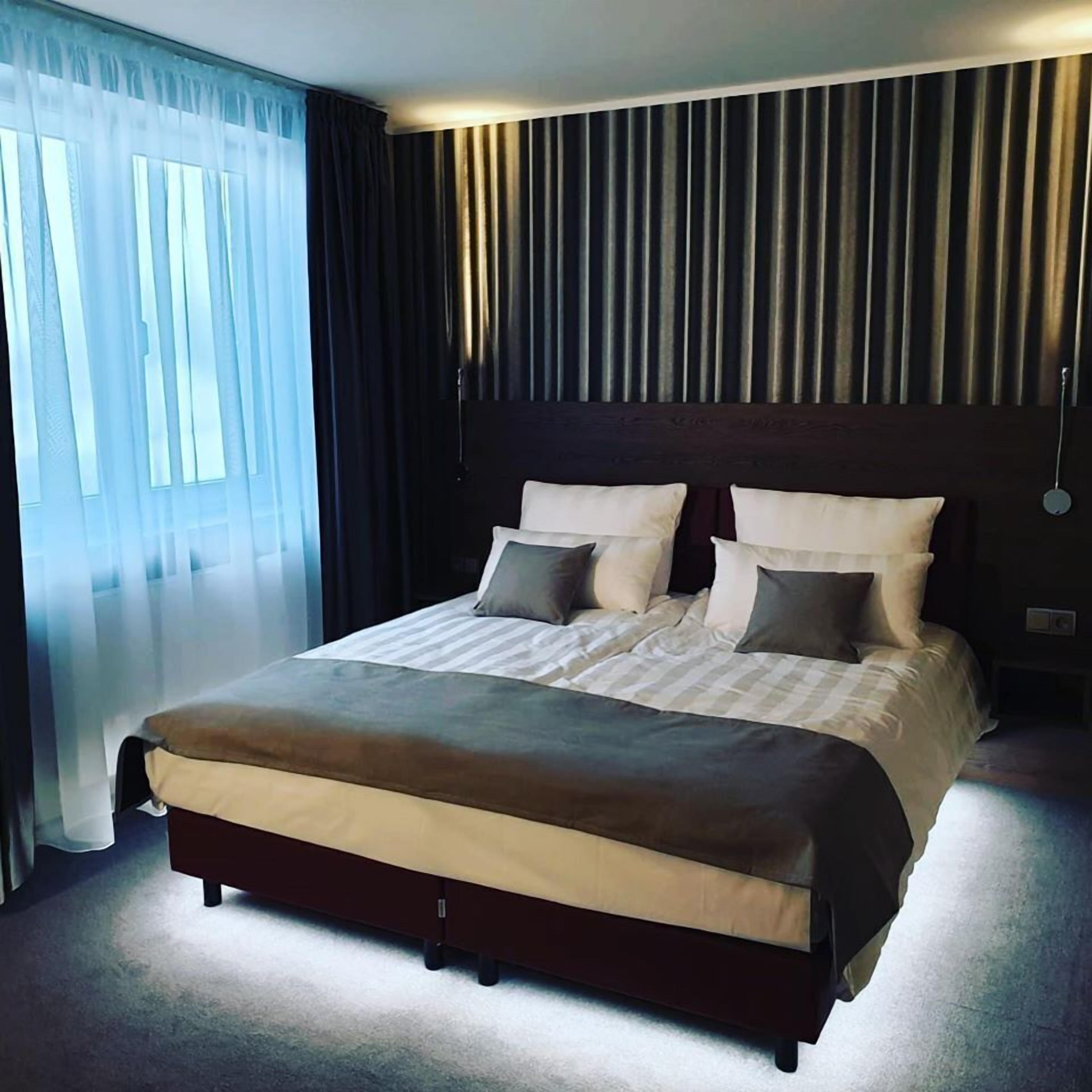 Una habitación de hotel moderna con una cama grande, cabecero oscuro y cortinas translúcidas en la ventana.