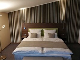 Una habitación de hotel con una cama grande, cabecera con dos almohadas, una mesita de noche y una lámpara de pie.