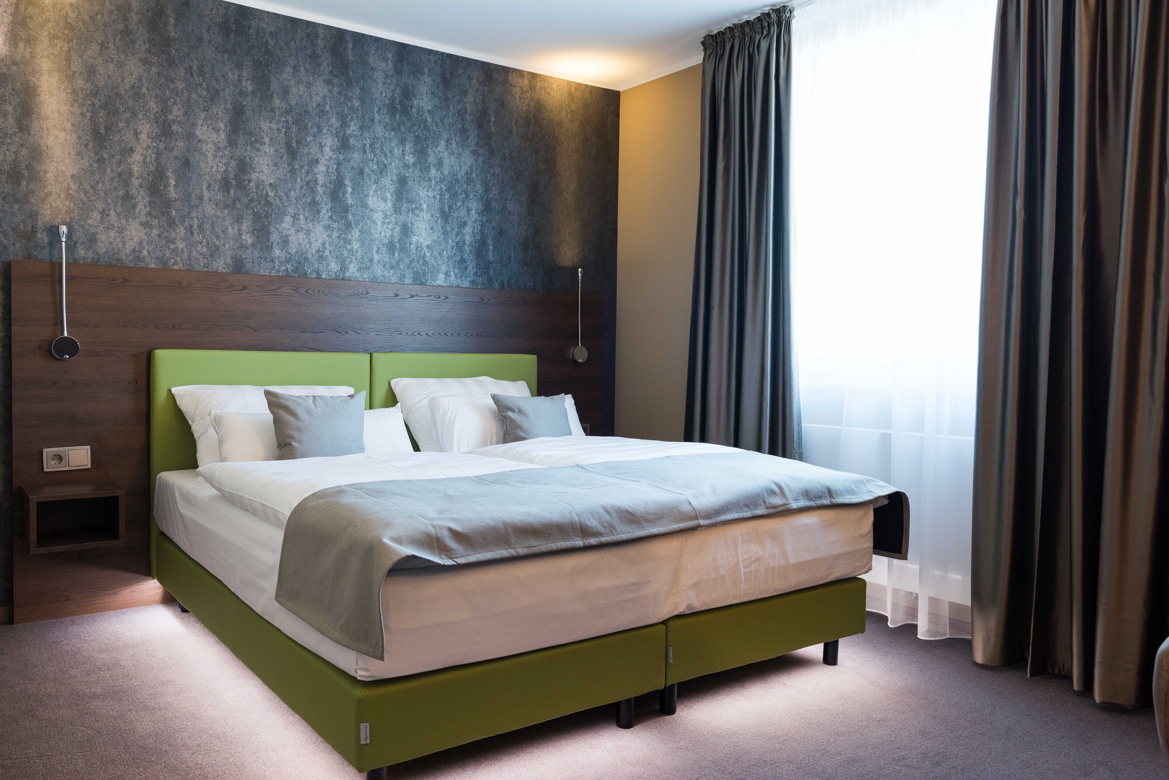 Una habitación de hotel moderna con una cama verde, un cabecero de madera, un ventanal con cortinas y una pared con textura.