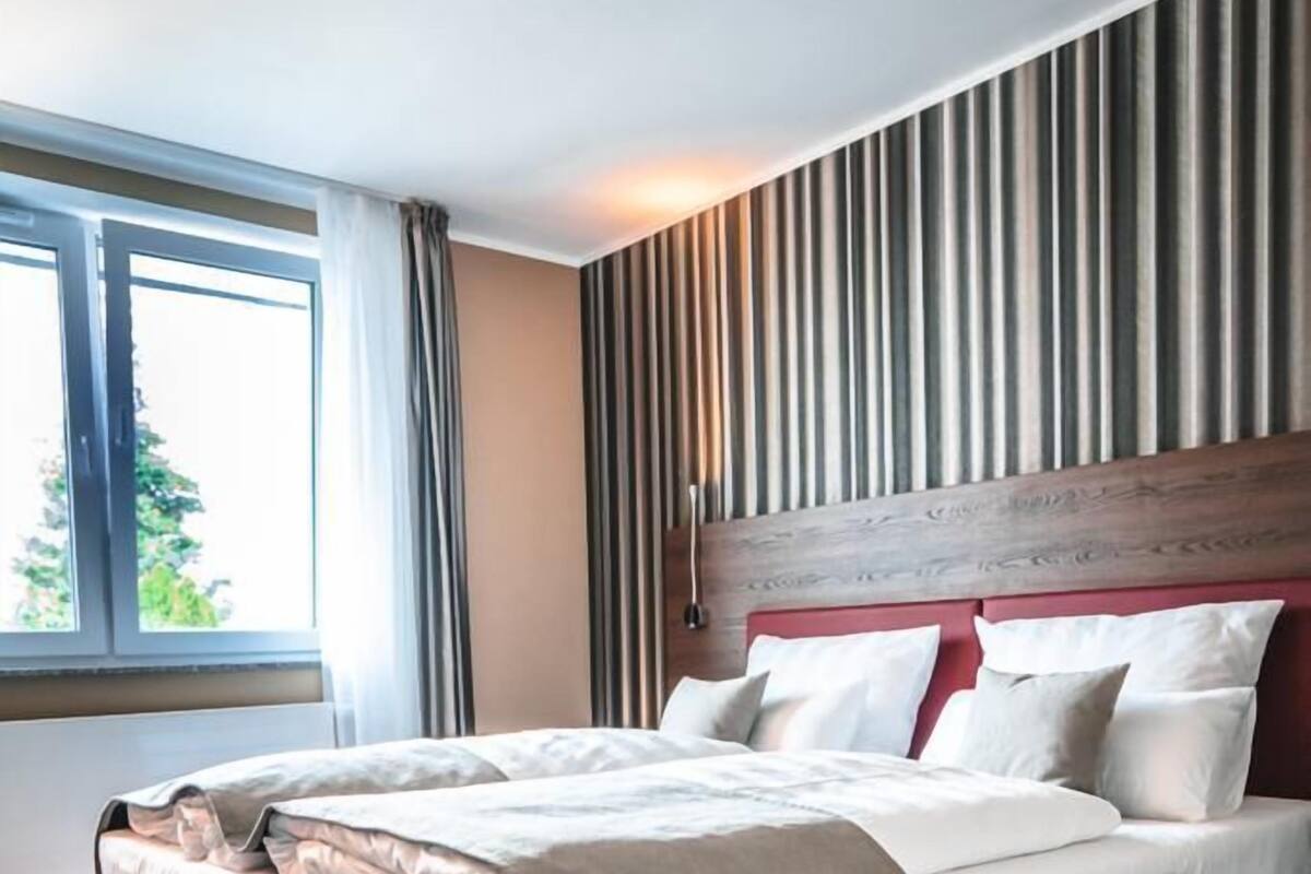 Een moderne hotelkamer met een groot bed, een houten hoofdeinde en een groot raam met gordijnen.