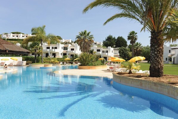 A swimming pool with sun loungers and umbrellas, surrounded by palm trees and white buildings.
