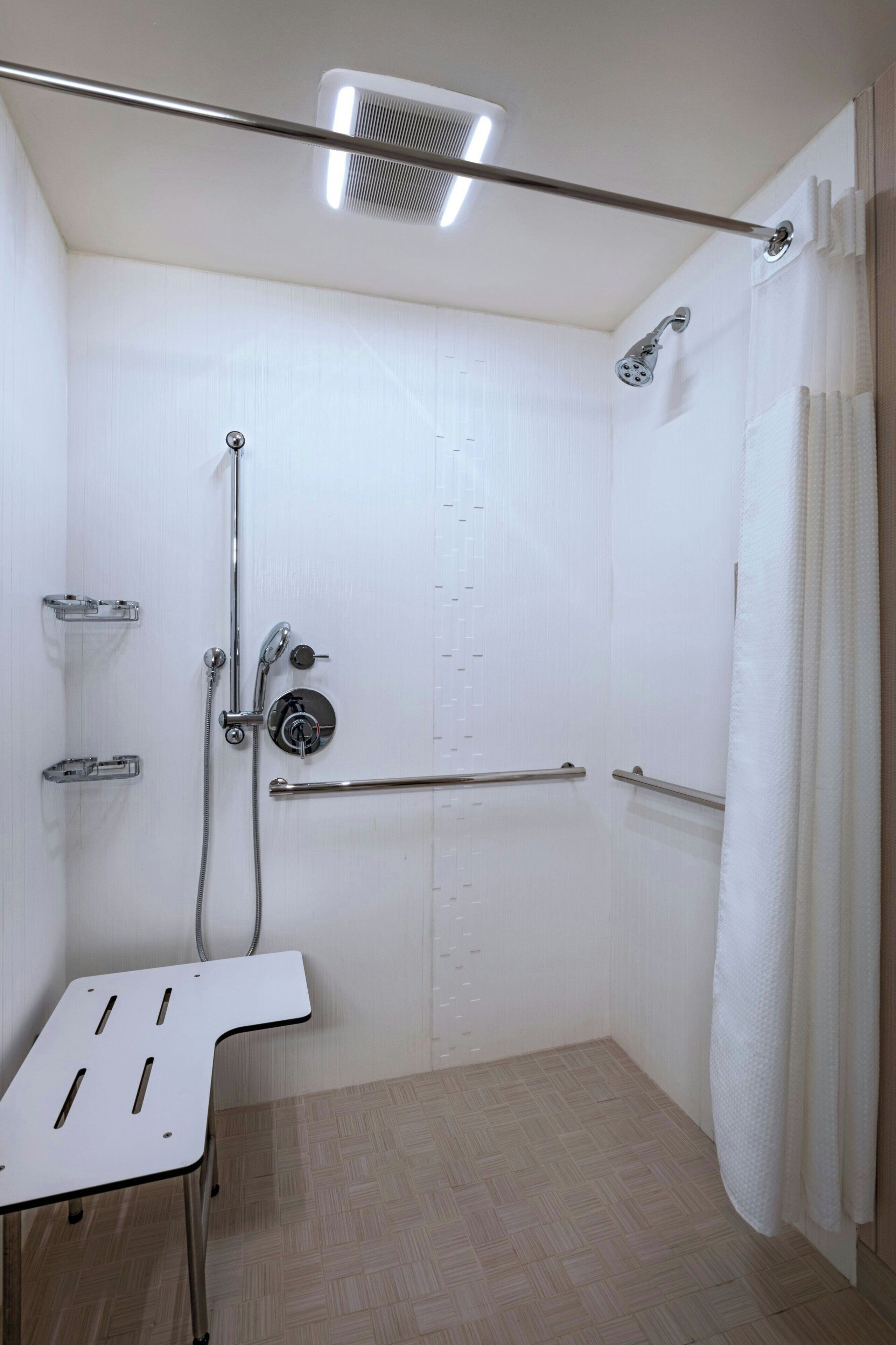 A small, white, walk-in shower with a grab bar, a handheld showerhead, and a ceiling-mounted light fixture.