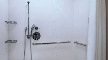 A small, white, walk-in shower with a grab bar, a handheld showerhead, and a ceiling-mounted light fixture.