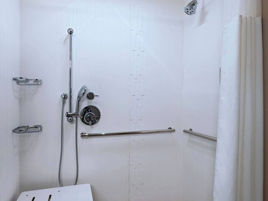 A small, white, walk-in shower with a grab bar, a handheld showerhead, and a ceiling-mounted light fixture.