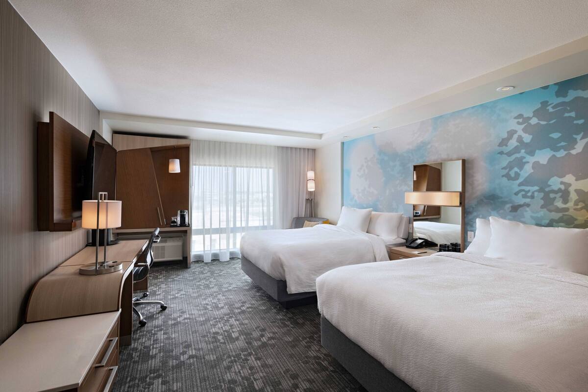 A hotel room with two beds, a desk, a chair, a lamp, a mirror, and a large window with curtains.