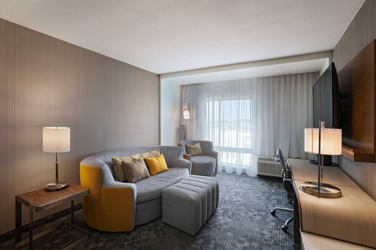 A modern hotel room with a sectional sofa, a small armchair, a desk with a chair, and a flat-screen TV.