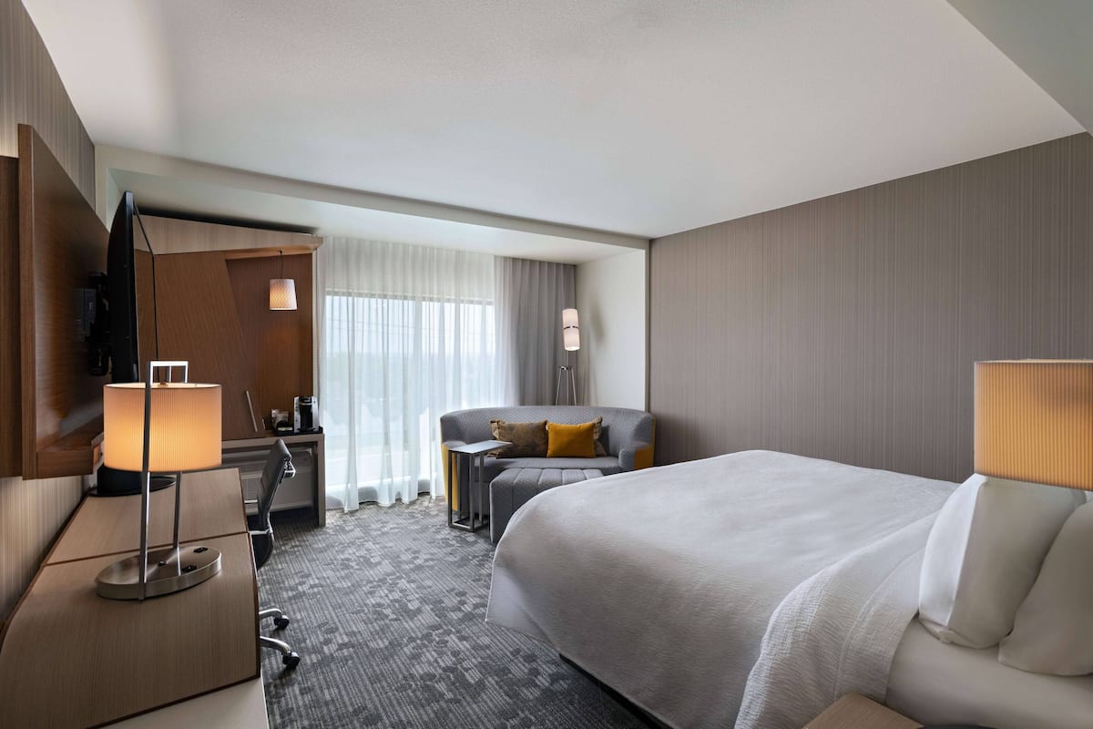 A hotel room with a large bed, a desk with a lamp, a chair, a sofa, and a TV.