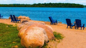 Una playa con rocas grandes, sillas azules y vistas a un lago con un bosque al fondo.