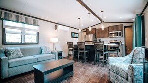 Una sala moderna con un sofá, una mesa de centro y un sillón estampado. Hay un comedor con una mesa y sillas, y una cocina con armarios y electrodomésticos.