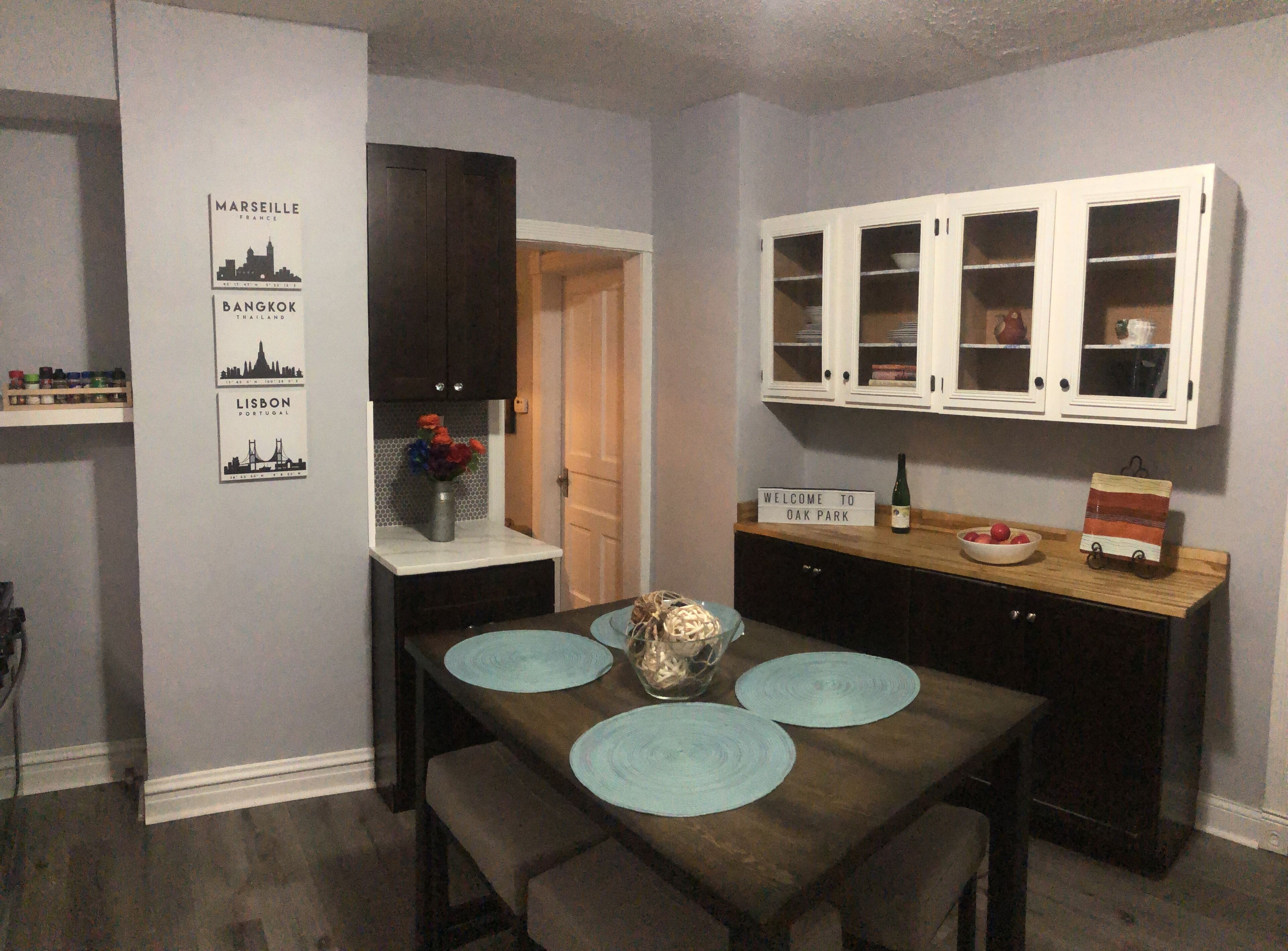 Un comedor con una mesa de madera, individuales azules y un jarrón de flores. Se observan armarios oscuros y un letrero que dice "Welcome to Oak Park".