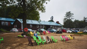 A beachside area with colorful Adirondack chairs, a large tree, and a building with a blue roof. - Great Blue Resorts - McCreary's Beach (Drummond-North Elmsley)