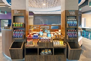 Exhibición de venta al público con diversos aperitivos y bebidas, incluyendo una sección de productos locales, con la sugerencia de solicitar artículos de conveniencia en la recepción.