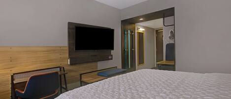 Una habitación de hotel con una cama grande, un televisor montado en la pared, una zona de estar con una silla y un espejo.