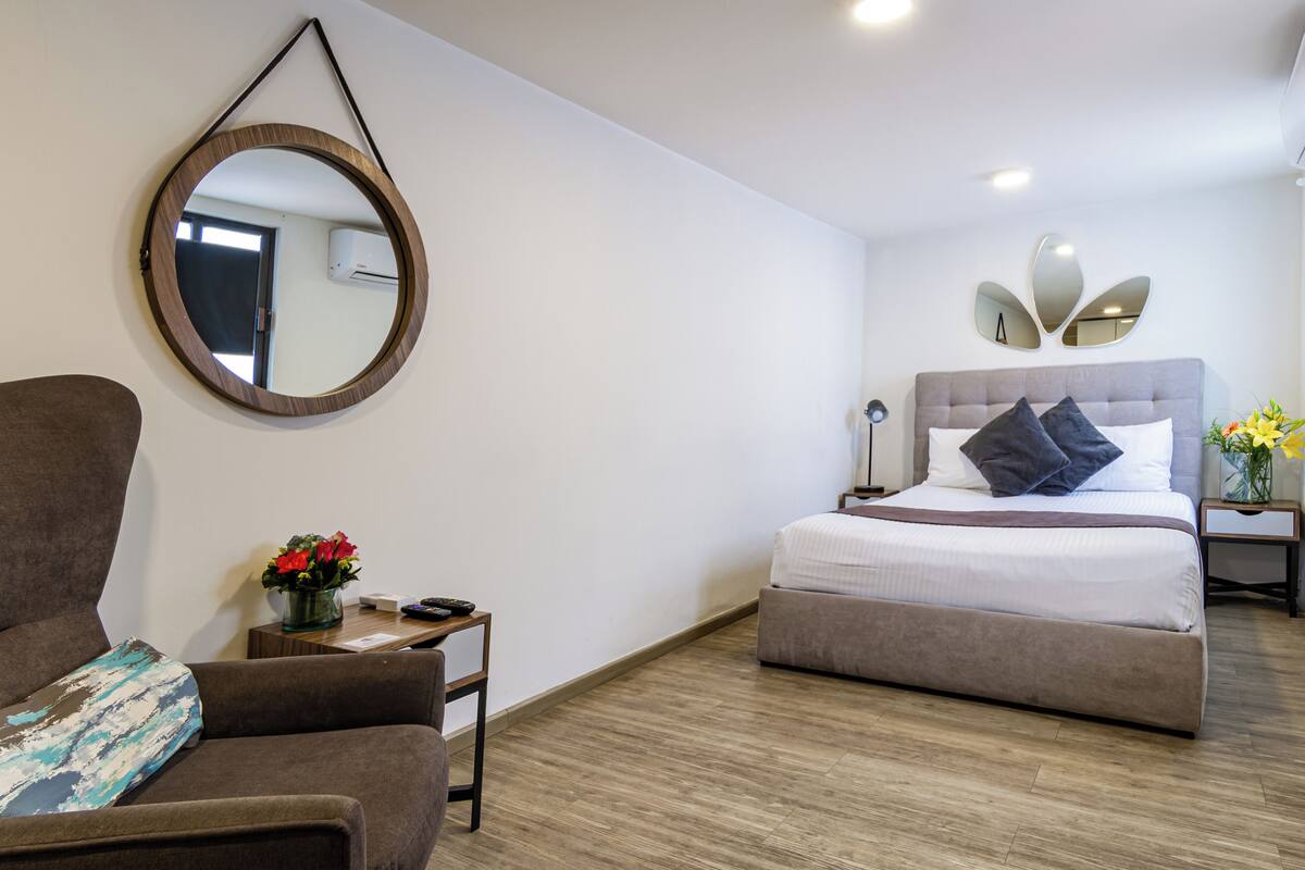 A bedroom with a bed, a chair, a small table with a vase of flowers, and a round mirror on the wall.