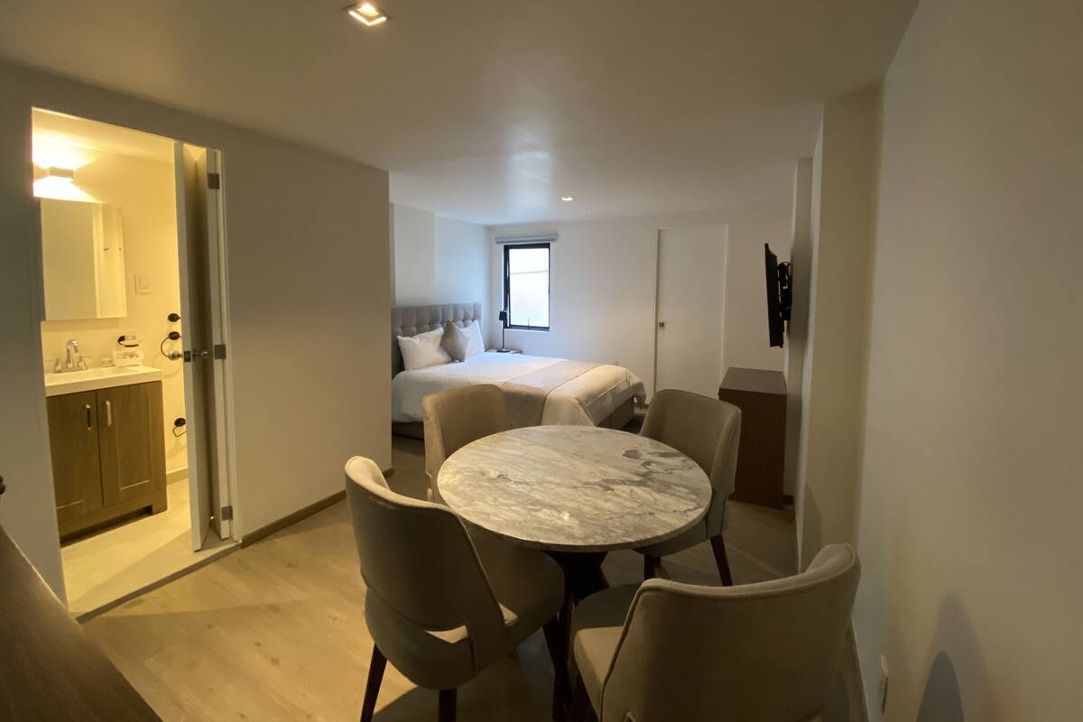 A modern hotel room with a bed, a dining table with chairs, a small kitchen area, and a bathroom.