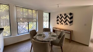 Un comedor con una mesa redonda, cuatro sillas y un banco. Se ven ventanas amplias con vistas a árboles y edificios.