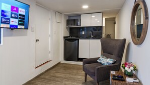 Una habitación de hotel moderna con una televisión de pantalla plana mostrando servicios de streaming, una kitchenette compacta, un sillón cómodo y una mesita con un jarrón de flores.