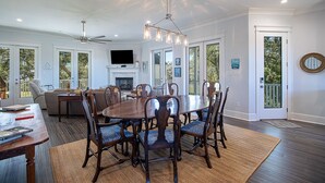 A spacious dining area with a large wooden table and chairs, a ceiling fan, and a mounted flat-screen TV. - EXPANSIVE PORCH-Gulf Breezes-Beach Views & Gorgeous Oak tree (Pass Christian)