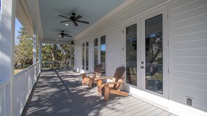 A covered porch with two wooden chairs and a table, white siding, and a ceiling fan. - EXPANSIVE PORCH-Gulf Breezes-Beach Views & Gorgeous Oak tree (Pass Christian)