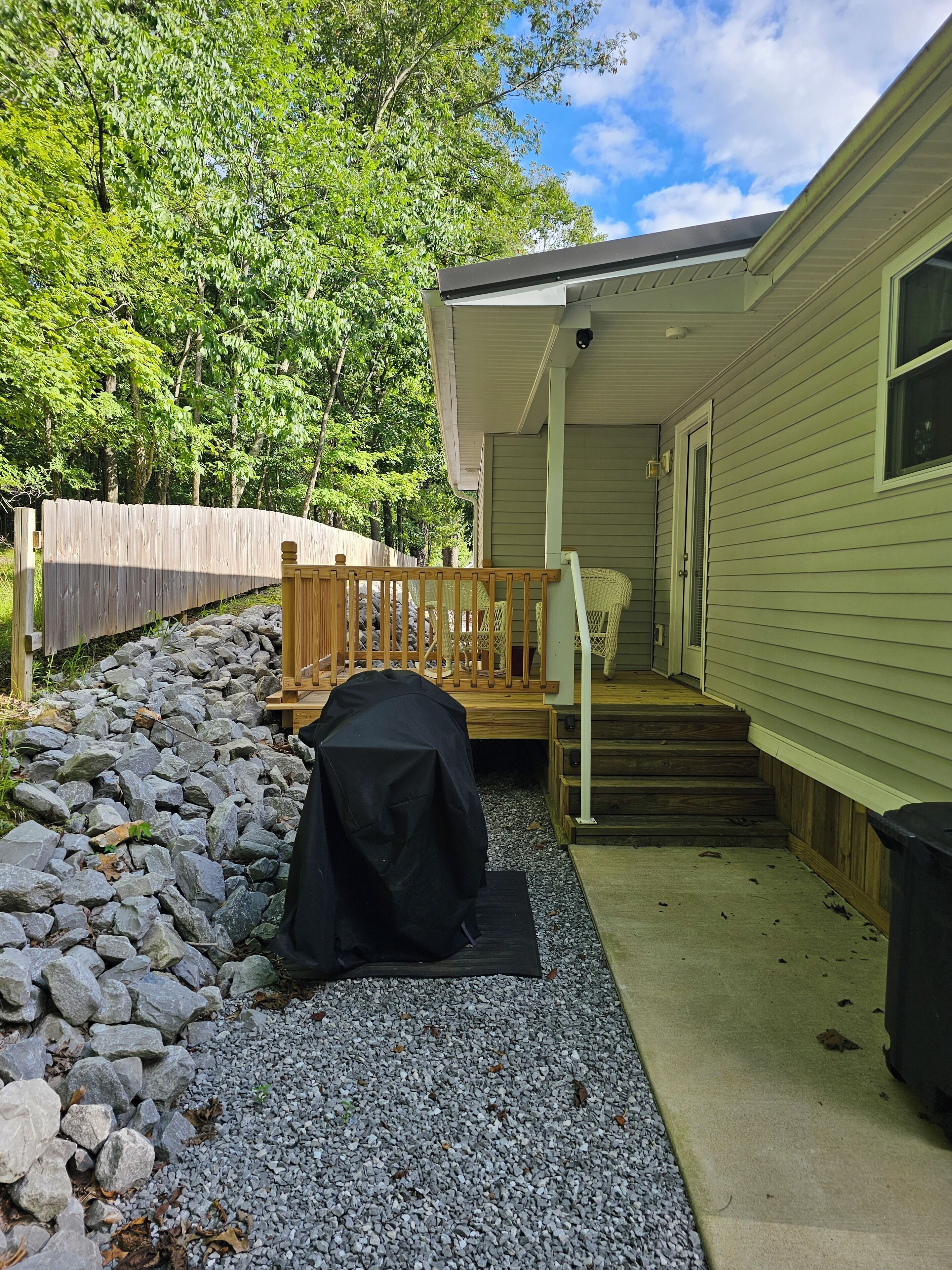 A covered outdoor area with a grill, a wooden deck, and a stone pathway leading to a house with a porch and steps.