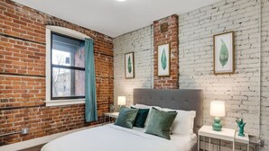 A bedroom with a brick wall, a large bed with green pillows, a window with a blue curtain, and framed leaf artwork on the wall. - Bright Event Perfect 4BDRM LOFT 7Min To Downtown (Detroit)
