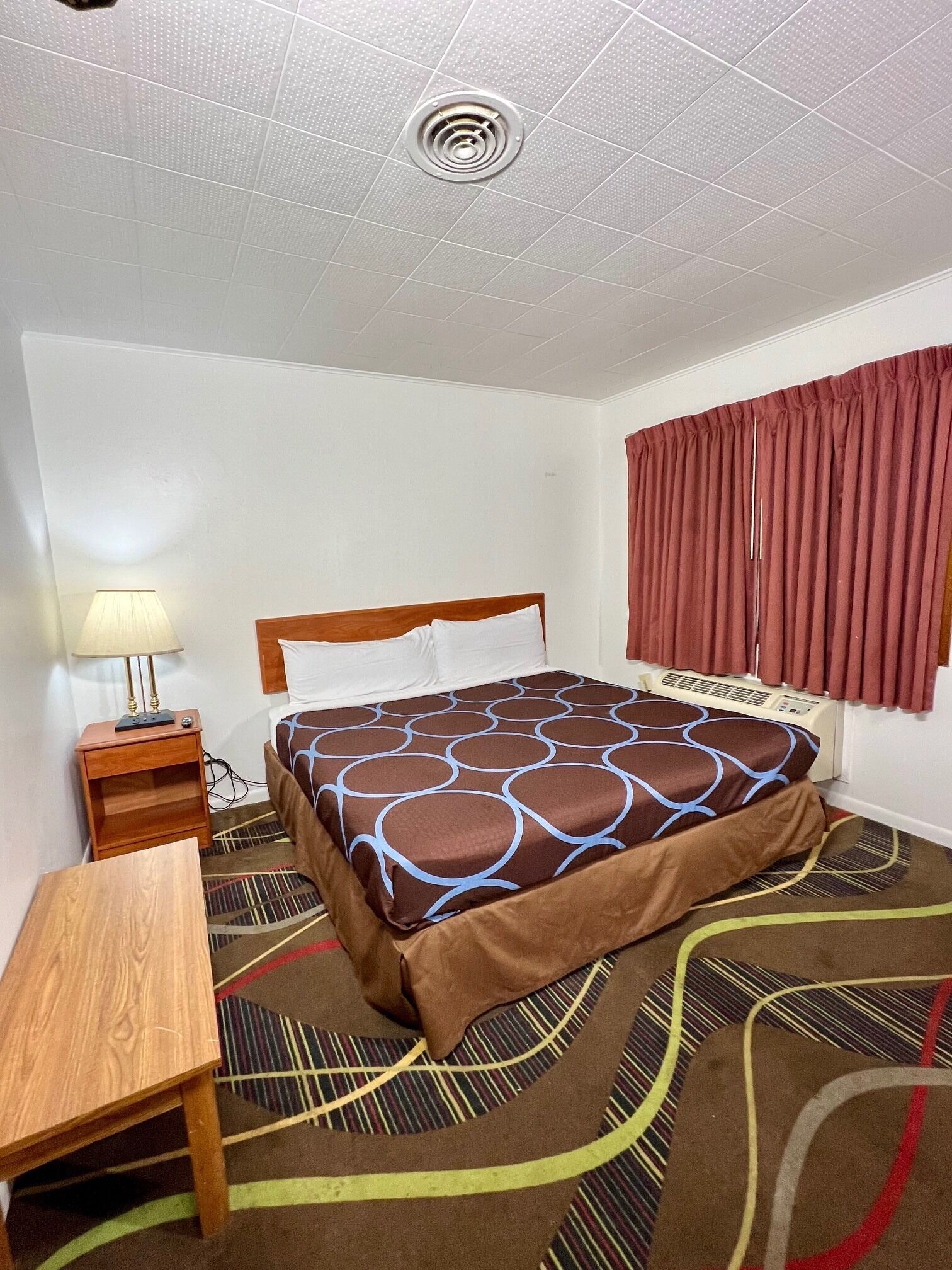 Ein Hotelzimmer mit Bett, Nachttisch, Lampe, Fenster mit Vorhängen und Lüftungsgitter an der Decke.