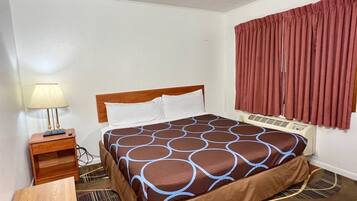 Ein Hotelzimmer mit Bett, Nachttisch, Lampe, Fenster mit Vorhängen und Lüftungsgitter an der Decke.
