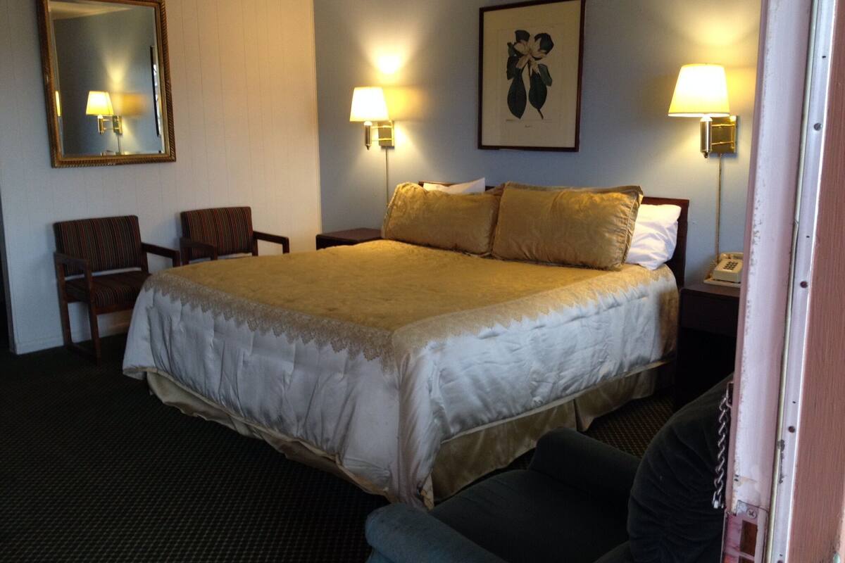 A hotel room with a large bed, two bedside lamps, a chair, a framed picture on the wall, and a telephone on the nightstand.