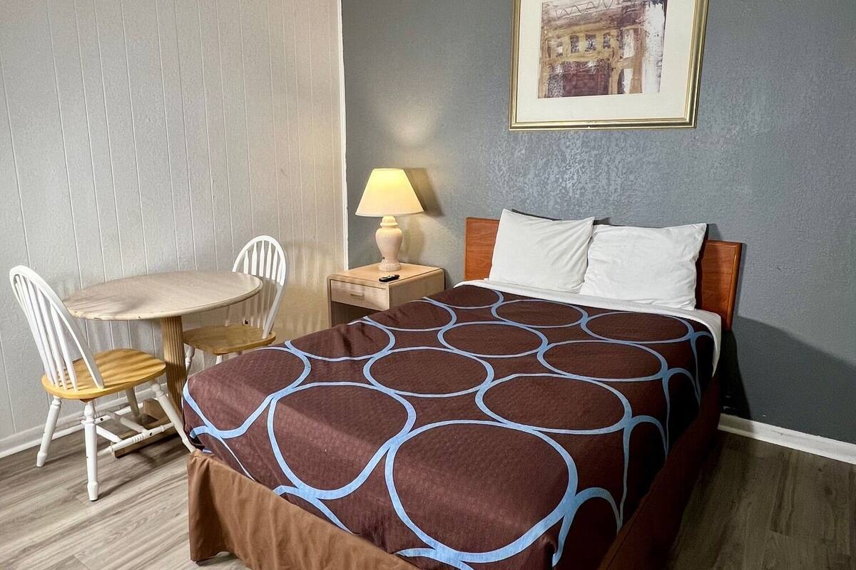 A hotel room with a bed, a desk, a chair, and a nightstand with a lamp.