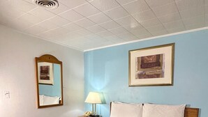 A hotel room with a bed, a nightstand, a lamp, a mirror, and a framed picture on the wall. - Village Inn (Salina)
